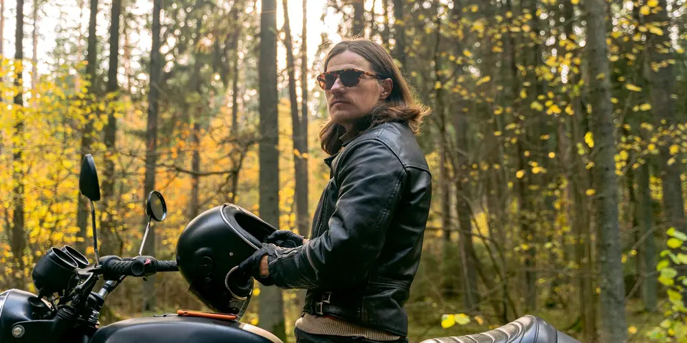 Person wearing a black leather jacket and sunglasses stands beside a motorcycle in a sunlit forest.