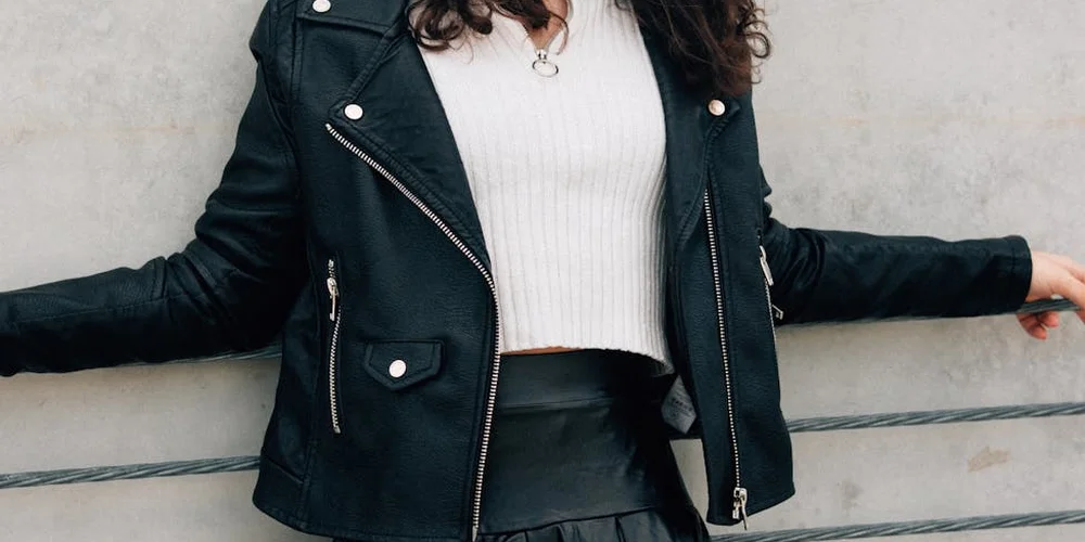 A black leather jacket is worn by a person standing against a light concrete wall, arms extended.