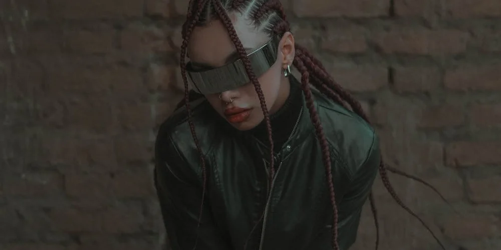 Person wearing a dark green leather jacket and protective eyewear with braided hair, standing against a brick wall.