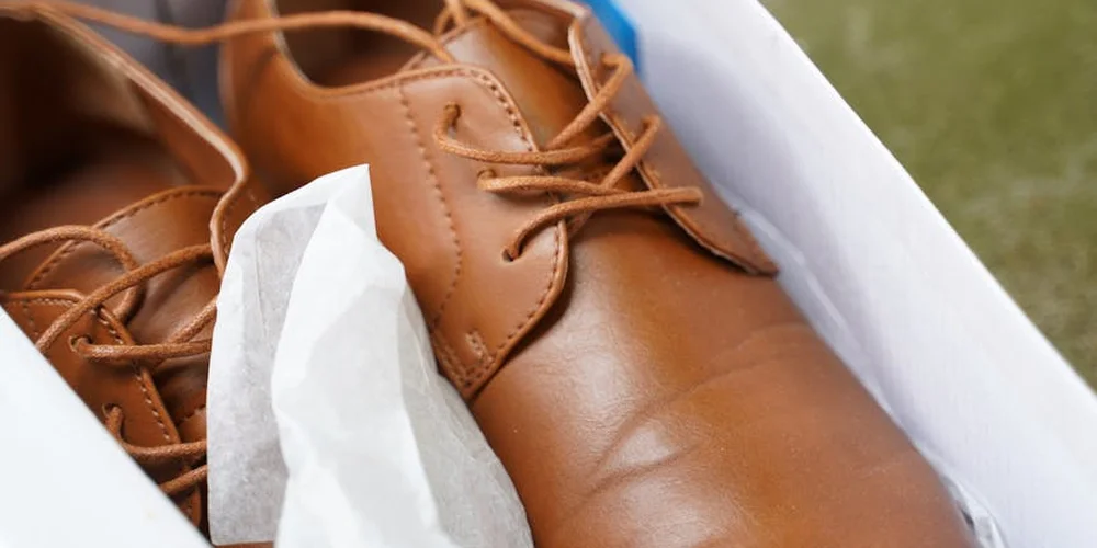 Brown leather shoes tucked in a white box with tissue paper, illustrating careful storage.
