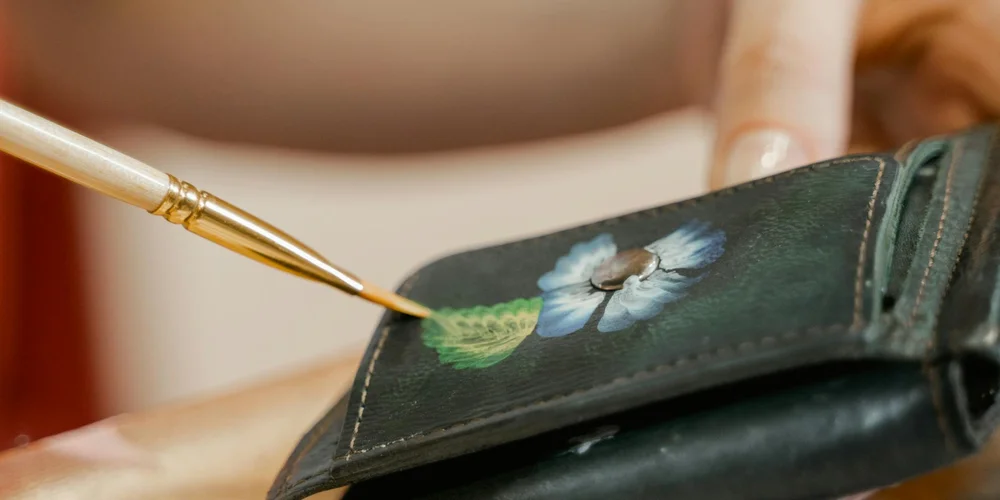 Close-up of a hand painting a blue flower onto dark leather with a fine brush, illustrating prep work for leather painting.