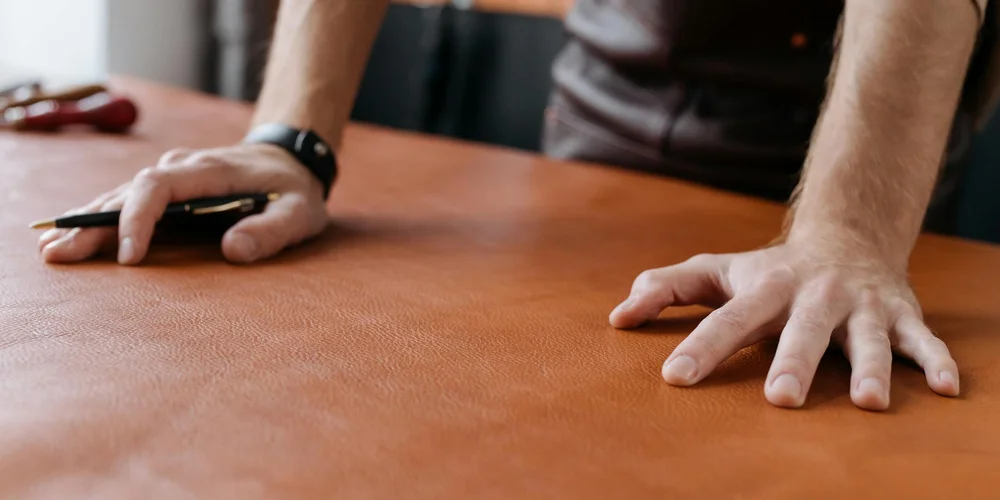 Close-up of a brown leather surface with a person's hands resting on it; one hand holds a pen, suggesting inspection of patina development.