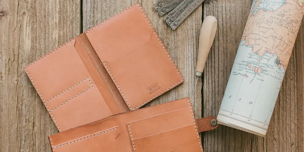 Two tan leather wallets on a weathered wooden surface with visible stitching, a rolled map, and a wooden-handled tool, highlighting the texture and finish of fine leather.