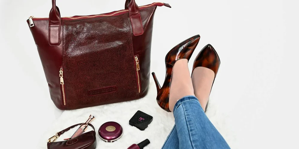 A burgundy leather handbag with makeup items and brown high-heeled shoes on a white background