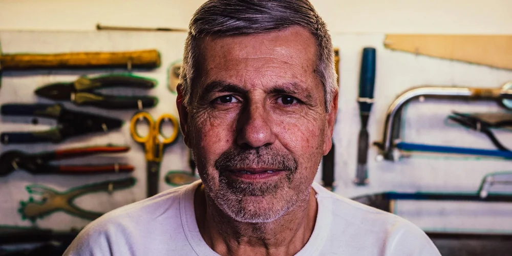 A middle-aged craftsman with short gray hair and stubble stands in a workshop, with leatherworking tools hung on the wall behind him.