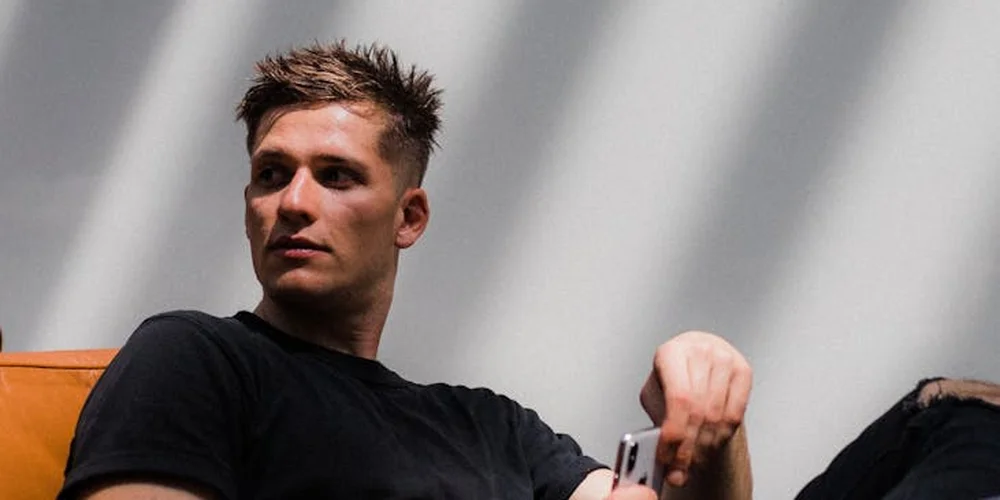 Man in a black t-shirt sitting on an orange upholstered chair, holding a tool and looking to the side against a neutral backdrop.