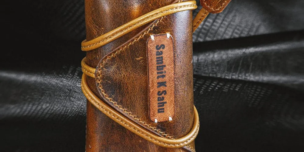 Close-up of a brown leather item featuring textured leather, stitching, and a vertical leather tag with straps.