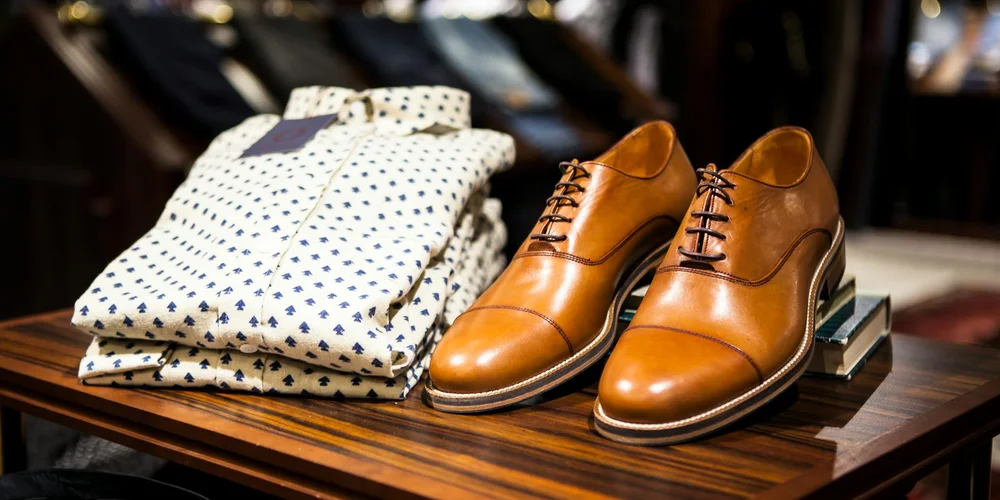 Brown leather dress shoes and folded patterned shirts on a wooden table