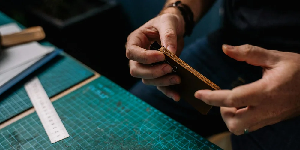 Close-up of hands measuring a leather strip on a green cutting mat with a ruler, illustrating careful leather handling.