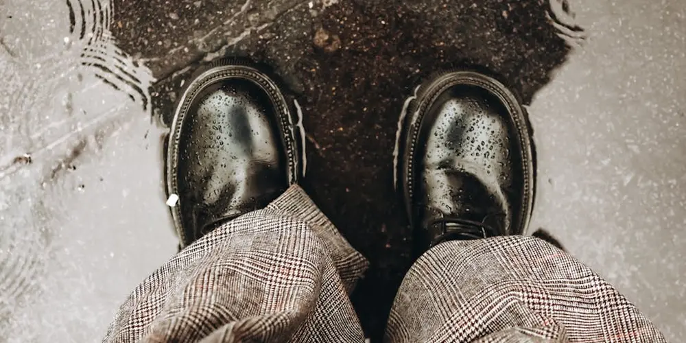 View of black leather boots standing on a damp, reflective surface with plaid pants visible, illustrating leather care in wet conditions.