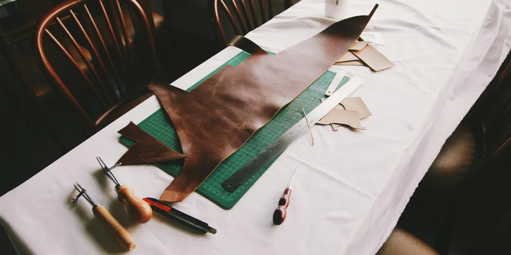 Brown leather pieces arranged on a green cutting mat with knives, a ruler, and small scraps on a white workshop table.
