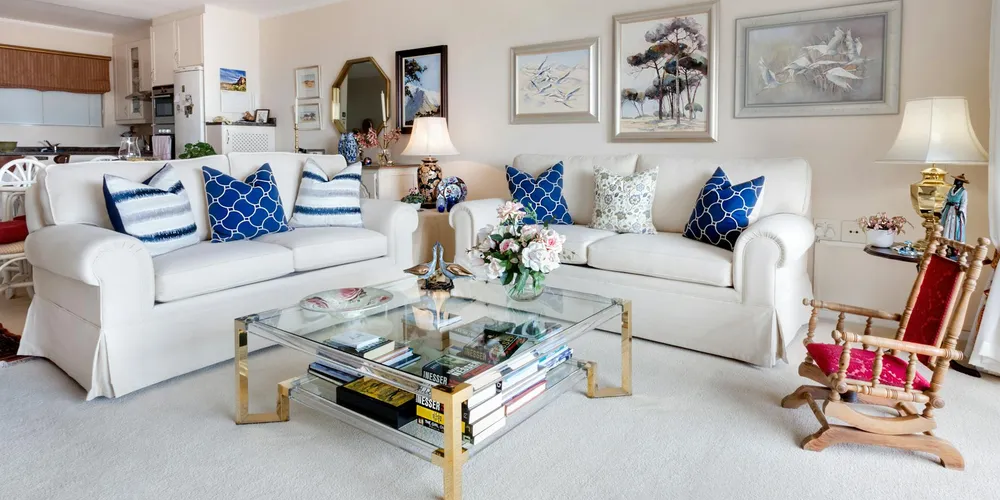Bright living room with white fabric sofas, blue patterned throw pillows, a glass coffee table, and framed artwork on light-colored walls.
