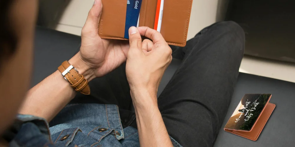 A person wearing casual clothes holds a brown luxury leather wallet, with additional wallets nearby and a brown leather watch on the wrist.