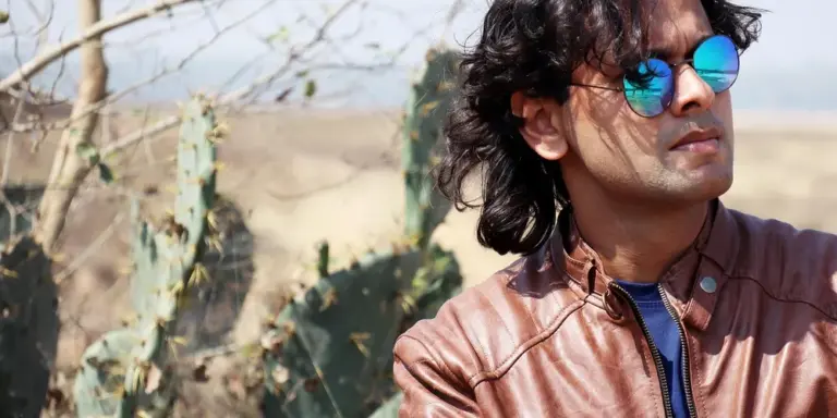 Man wearing a brown leather jacket and blue sunglasses standing outdoors in a desert-like setting.