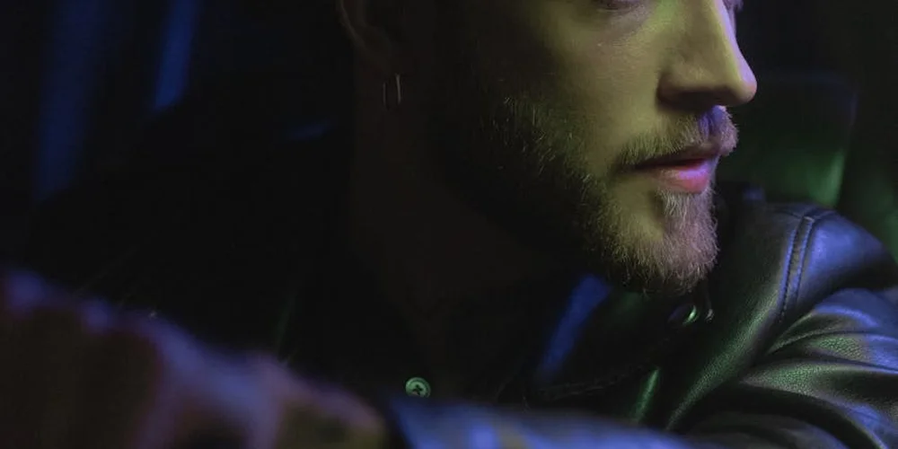 Close-up profile of a man wearing a black leather jacket in a moody, dimly lit setting.