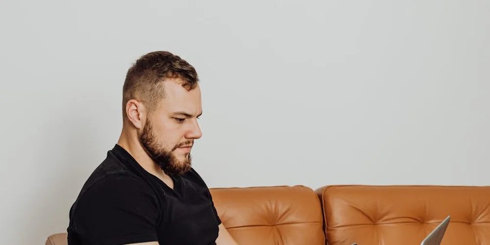Man with a beard wearing a black shirt sits on a tan leather sofa, looking to the side.