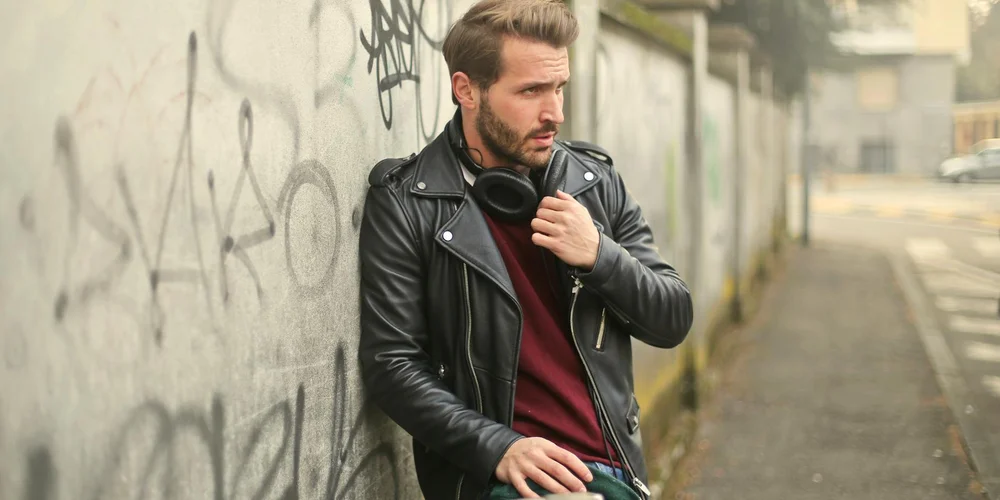 A man wearing a black leather jacket and burgundy sweater stands beside a graffiti-covered wall on a city sidewalk.
