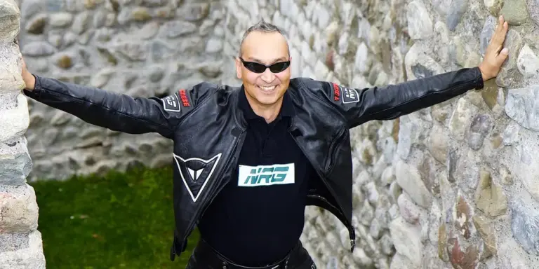 Model in a black leather jacket with patches wearing sunglasses, standing between stone walls with arms outstretched.