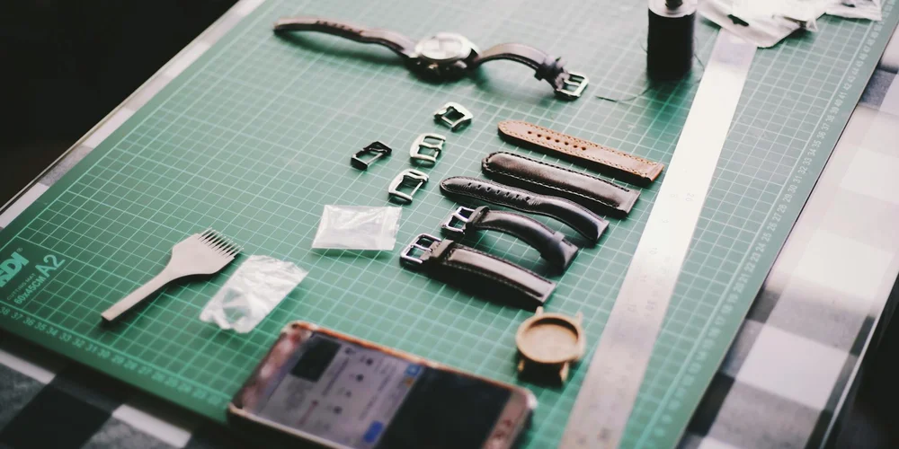Leather care tools and hardware laid out on a green cutting mat, including buckles, straps, a brush, and a ruler, illustrating how to select the appropriate care for different types of leather.