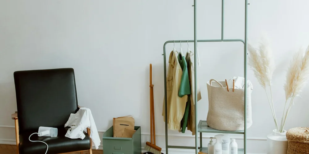 Minimalist room with a metal clothing rack holding jackets and a bag, a chair with a cloth, a broom, and decorative dried plants, suggesting a storage area where leather items might be cleaned or evaluated.