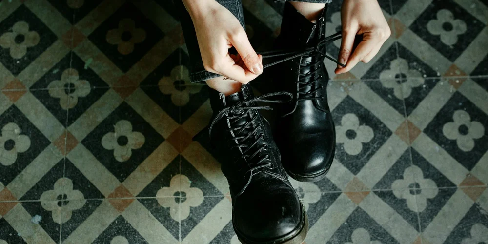 Close-up of hands tying the laces on black leather boots, set against a patterned tile floor.