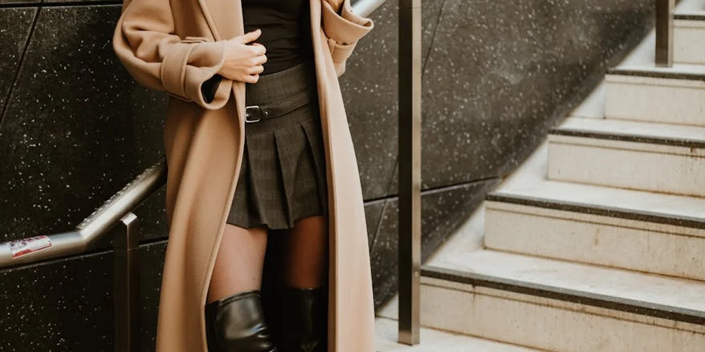 Person wearing a tan coat and black outfit standing next to stairs, wearing knee-high black leather boots.