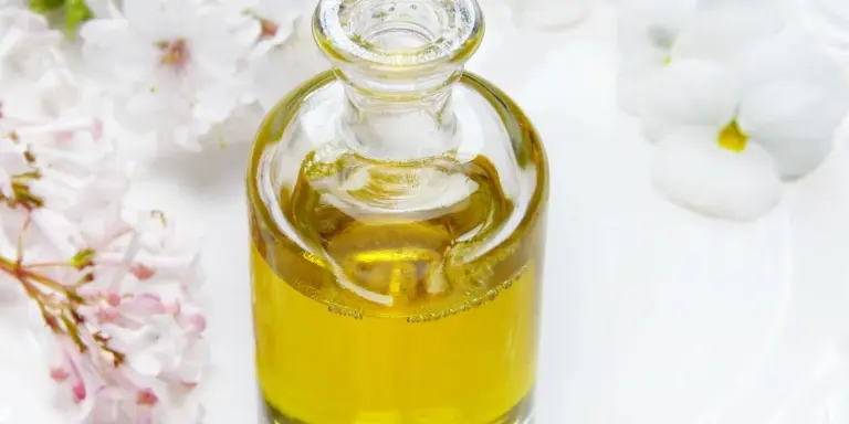 Glass bottle of golden mink oil on a white surface, with soft pink and white flowers in the background.