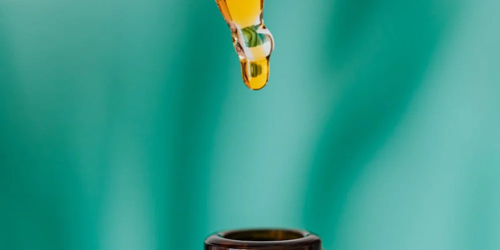 Close-up of a glass dropper releasing a golden drop of oil with a soft green background, representing mink oil used for leather conditioning.