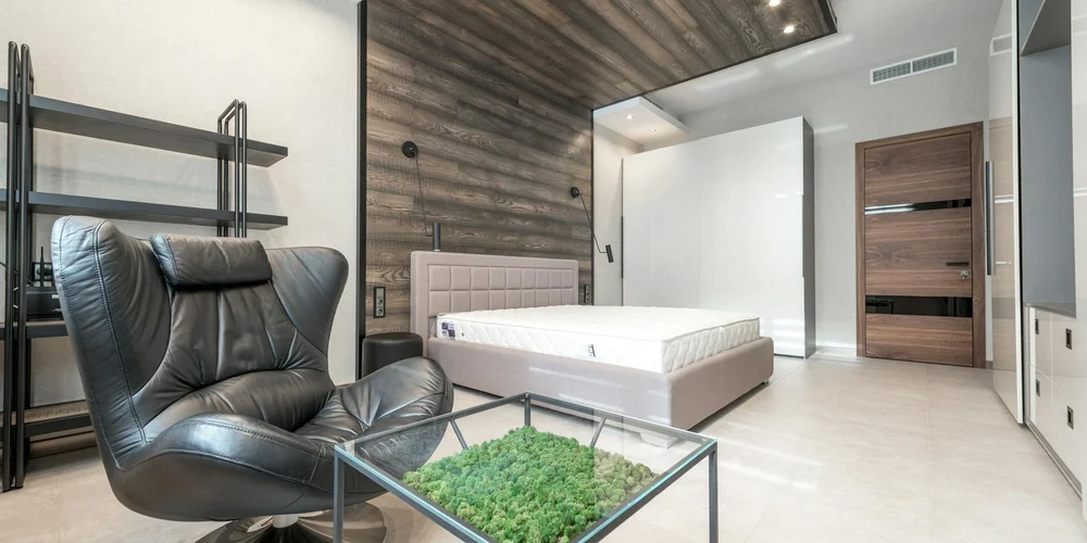 A contemporary room features a black leather lounge chair beside a glass coffee table and a bed with a tufted headboard, with wood paneling and neutral decor.