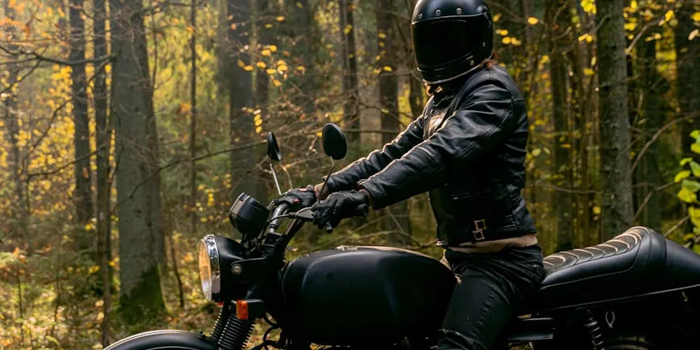 A motorcyclist wearing full protective leather gear sits on a black motorcycle in a forest with fall foliage.