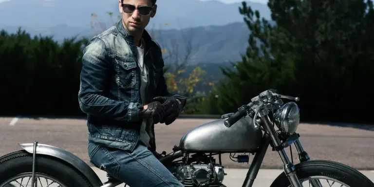 A motorcycle rider wearing a blue denim jacket and gloves stands beside a silver motorcycle on an open road, with trees and mountains in the background.