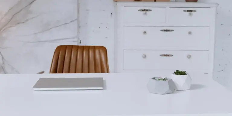 Brown natural leather chair at a white desk with a notebook and geometric planters, against a white marble wall and a white dresser in the background.