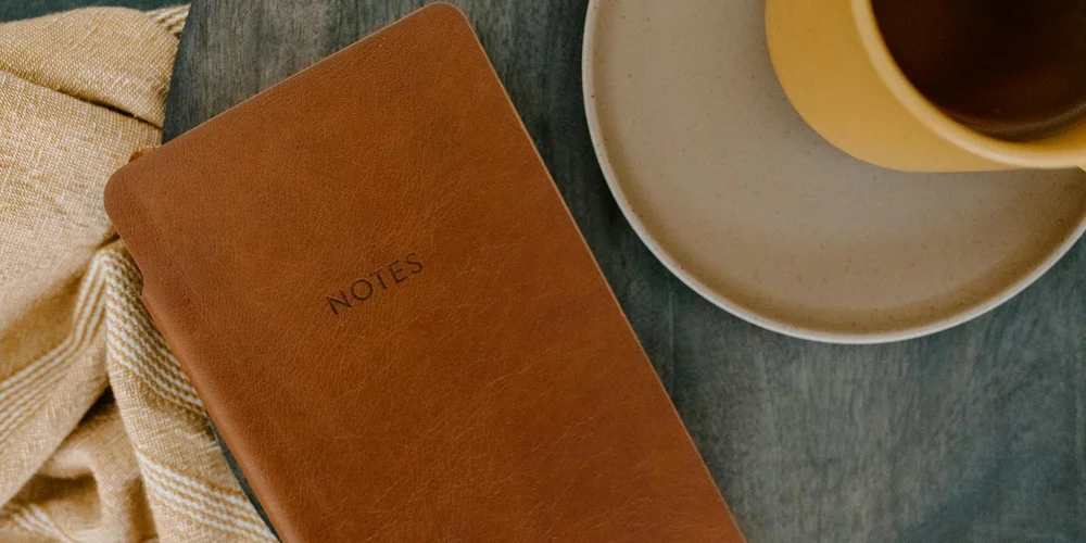 Tan leather notebook labeled NOTES on a dark wooden table, with a beige cloth and a cup of coffee on a saucer nearby.