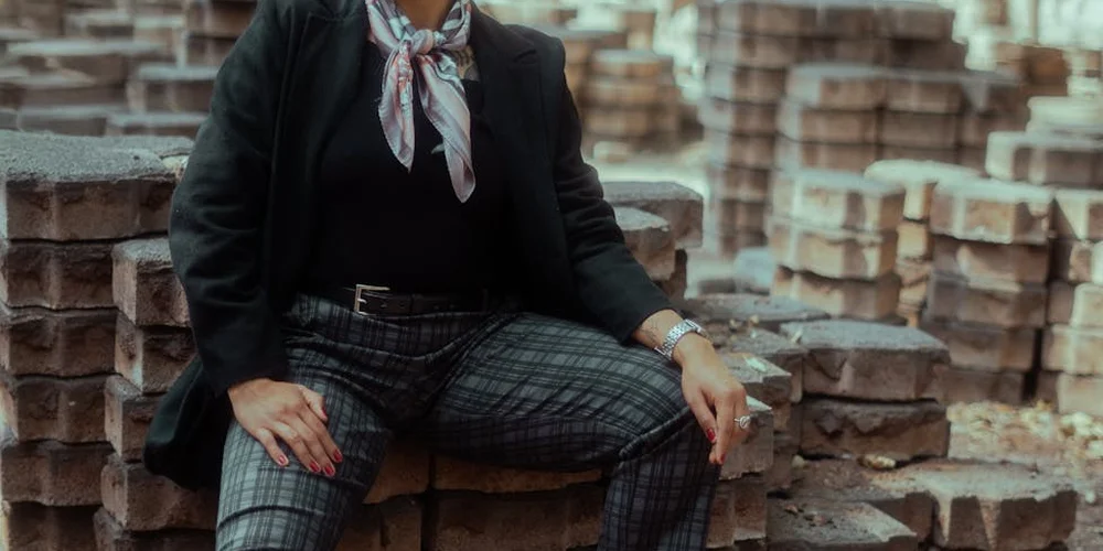 Person seated on stacked bricks wearing a black blazer, scarf, and plaid pants, illustrating a casual setting for discussing nubuck leather maintenance.