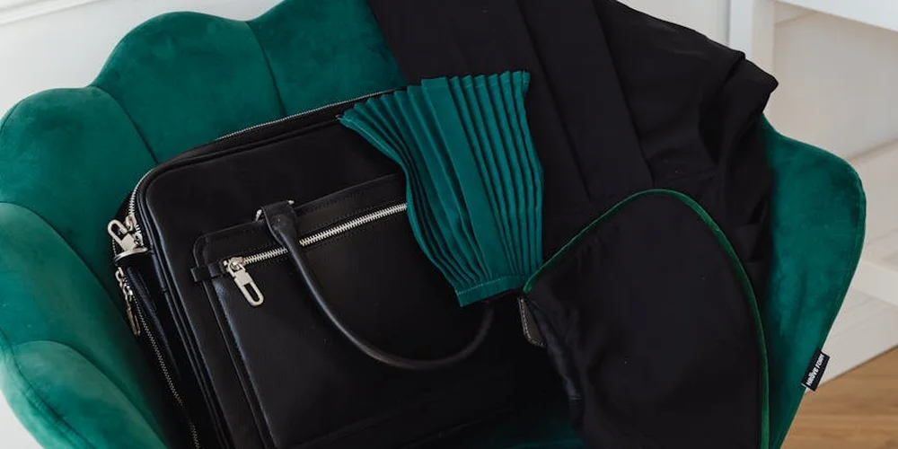 Two black leather bags on a green velvet chair; one appears to be patent leather with silver zippers, accompanied by a teal accessory nearby.