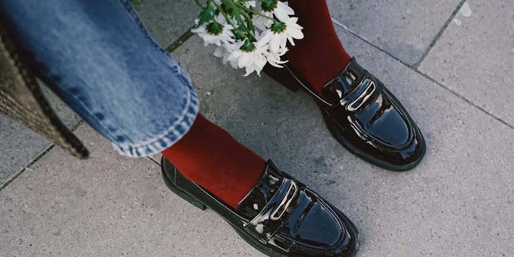 Top-down view of shiny black patent leather loafers on concrete pavement, worn with red socks and blue jeans, with a small bouquet of white flowers nearby.