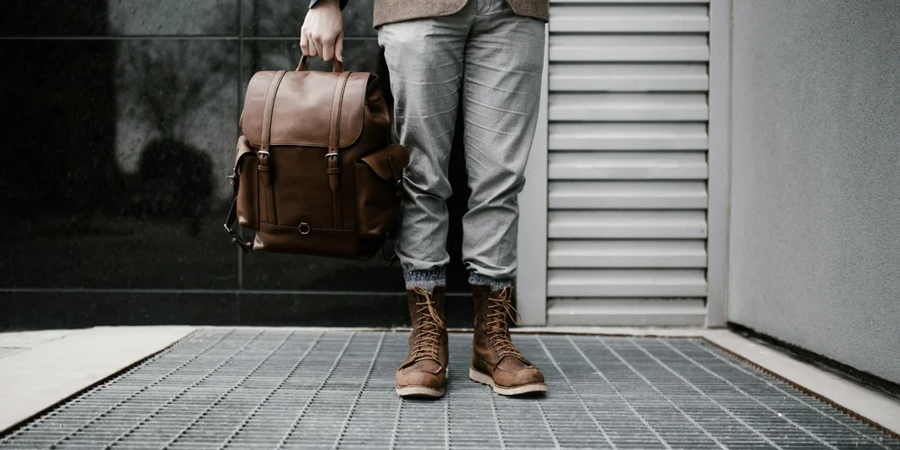 Person wearing gray jeans and brown leather boots holds a brown leather backpack outdoors.
