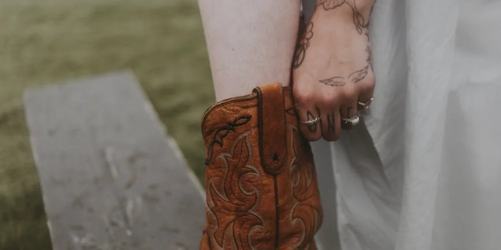 Brown embossed leather boot worn on the foot; a tattooed forearm and rings appear nearby in the image.