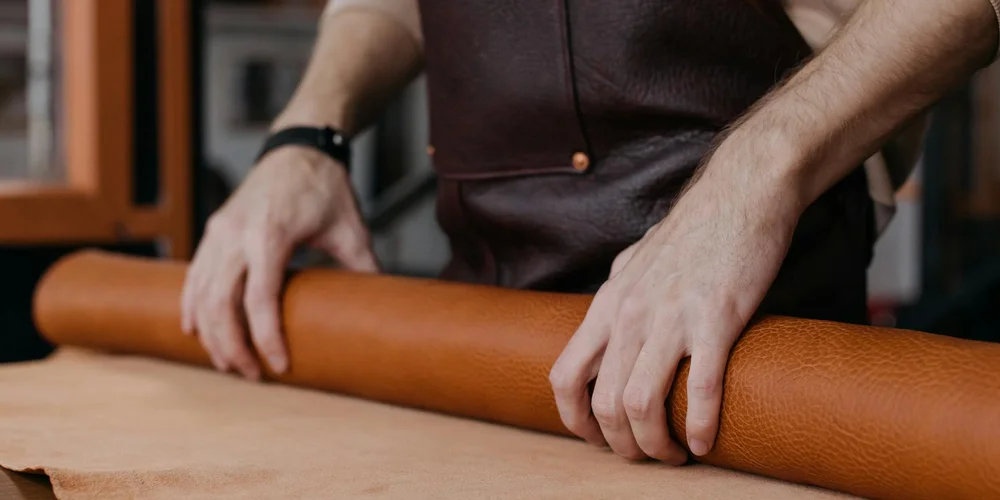 A craftsman smooths a long piece of brown leather on a workbench, preparing it for waterproofing.