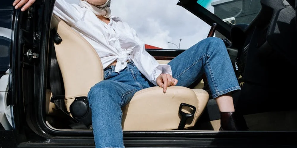 A person in blue jeans and a white shirt sits in the driver's seat of an open car, with the interior visible.