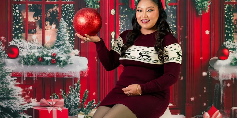 A fashion-forward woman in a burgundy knit dress poses with a red ornament in a festive Christmas setting, with a red door and holiday decor in the background.
