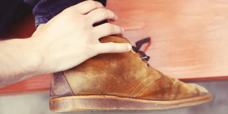 Close-up of a tan suede Puma sneaker being cleaned on a wooden surface, with a hand gently resting on the shoe.