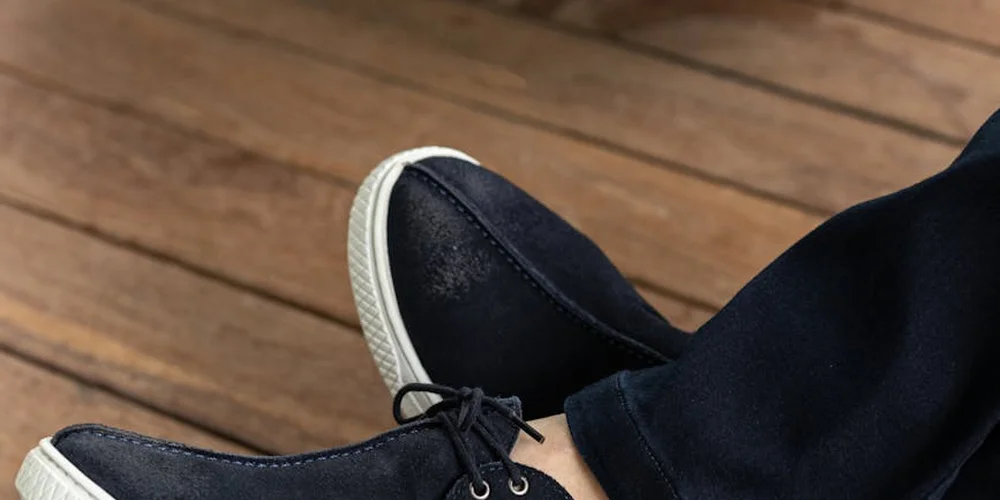 A pair of dark suede sneakers with white soles resting on a wooden floor.