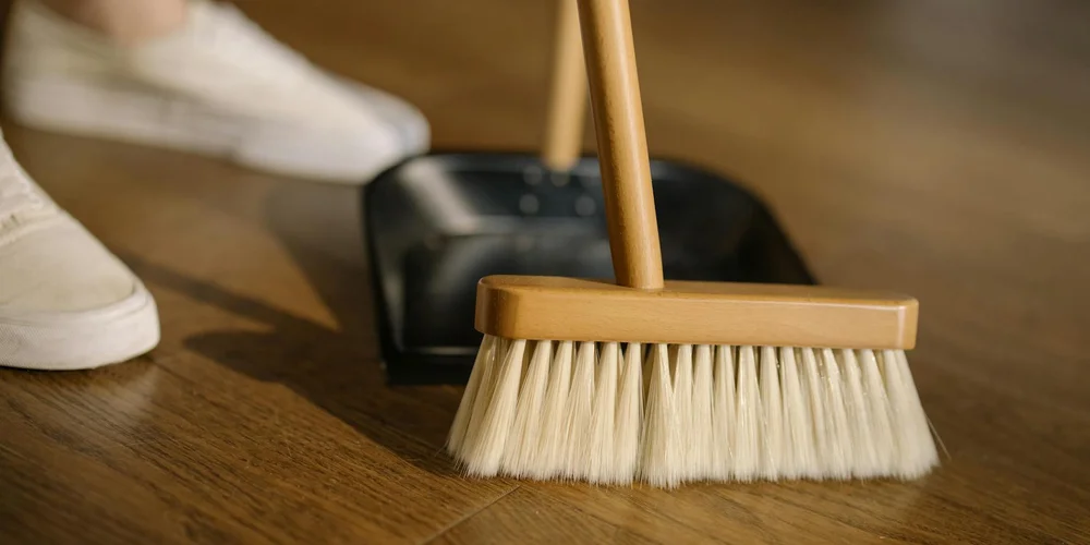 A wooden-handled cleaning brush rests on a wooden floor beside a black dustpan, with a white sneaker visible in the background.