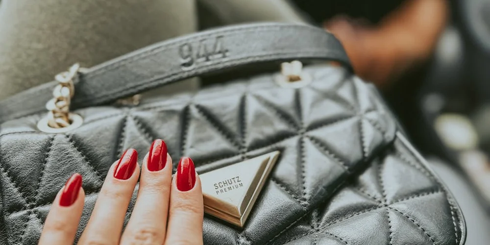 Close-up of a quilted black leather handbag with a chain strap and a hand with red-painted nails resting on its surface.