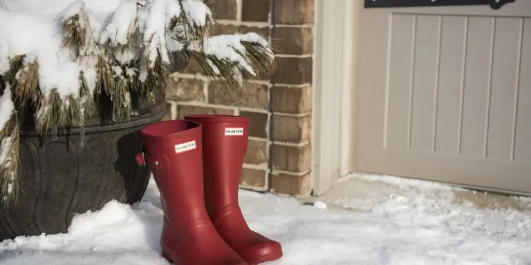 Red suede boots standing in the snow near a brick wall and a white garage door.