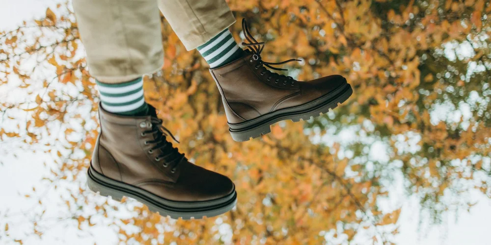 Brown leather boots with black soles and teal striped socks, suspended in midair against an autumn backdrop of orange and yellow leaves.
