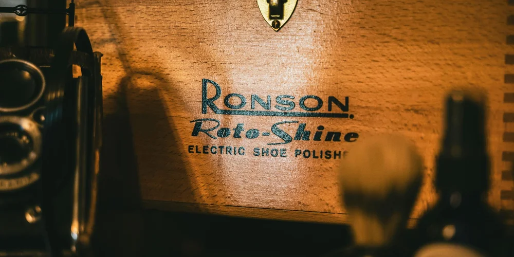 Close-up of a vintage wooden shoe polishing box labeled 'Ronson Rota-Shine' with a brush and an accessory bottle in the foreground.