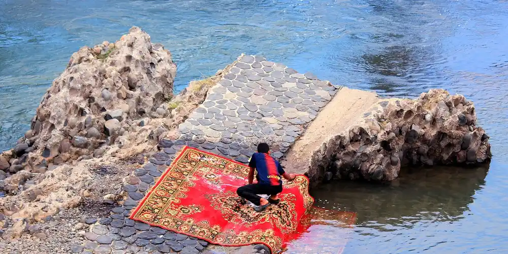 A person kneels on a red patterned rug laid over a rocky stone walkway beside a body of water.