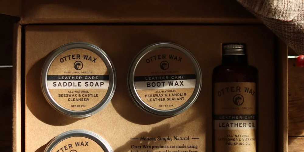 Close-up of Otter Wax leather care tins labeled Saddle Soap and Leather Wax, arranged in a wooden box with a cloth in view.